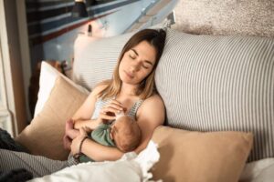 Young mother in striped top resting with newborn on couch, demonstrating the quiet moments of early motherhood that can be affected by postpartum depression. She appears contemplative while holding her baby in a naturally lit room with striped and beige pillows.